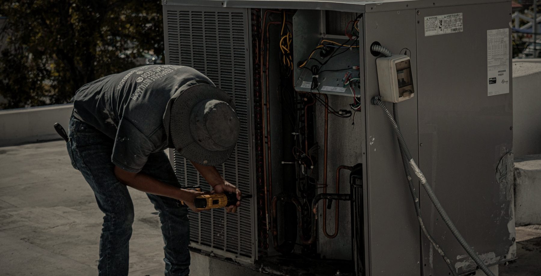 An HVAC technician servicing a rooftop condenser unit with a drill